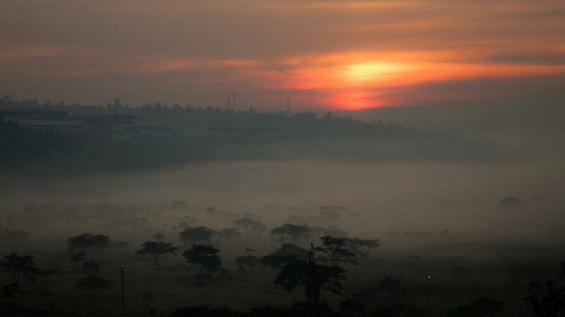 A glowing orange sun dips below the horizon over a misty, tree-filled valley in Kigali at dusk.