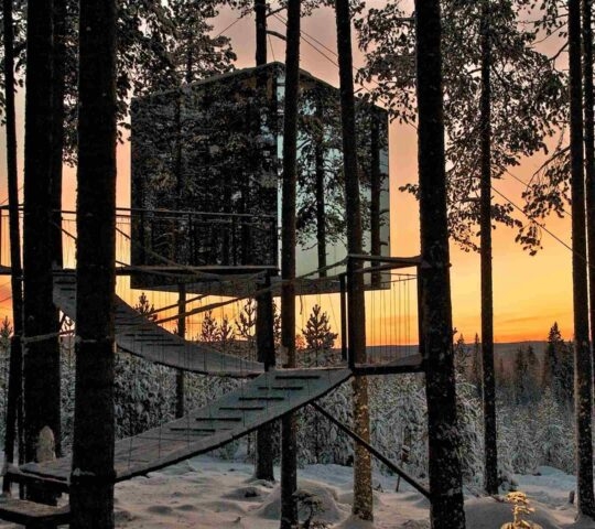 A square mirrored structure suspended in trees reflecting a snowy forest under a sunset sky.