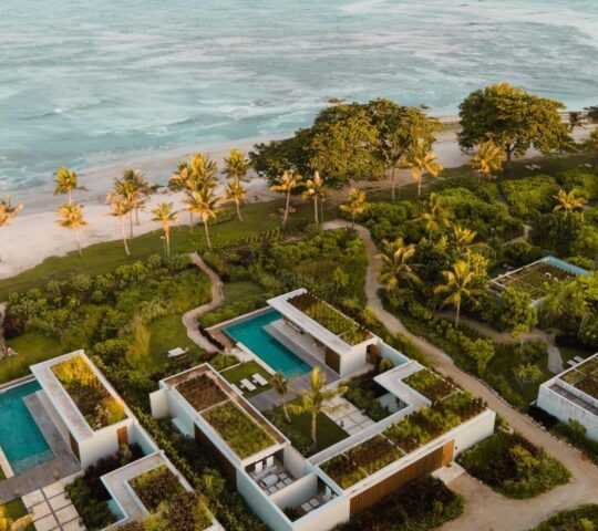 Aerial view of Cap Karoso's modern beachfront villas with green roofs and turquoise pools along a tropical coastline.