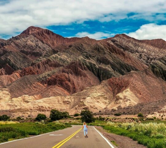 A paved road leads toward layered red and purple mountains with a person walking in the distance in Salta, Argentina.