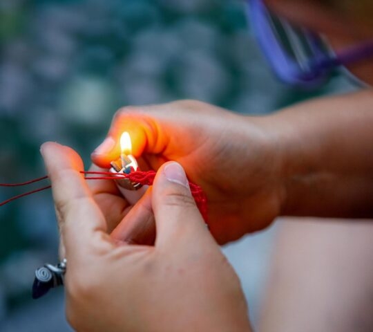 Close-up of an artisan's hands using a lighter to melt the ends of a red macramé string for a stone pendant.