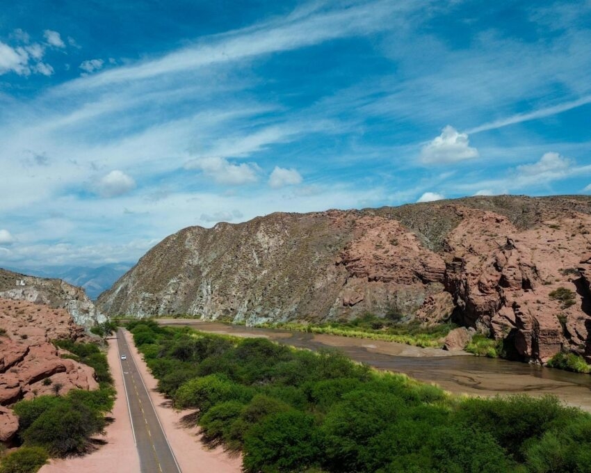 Aerial view of a paved road running through a red rock canyon and green valley under a bright blue sky.