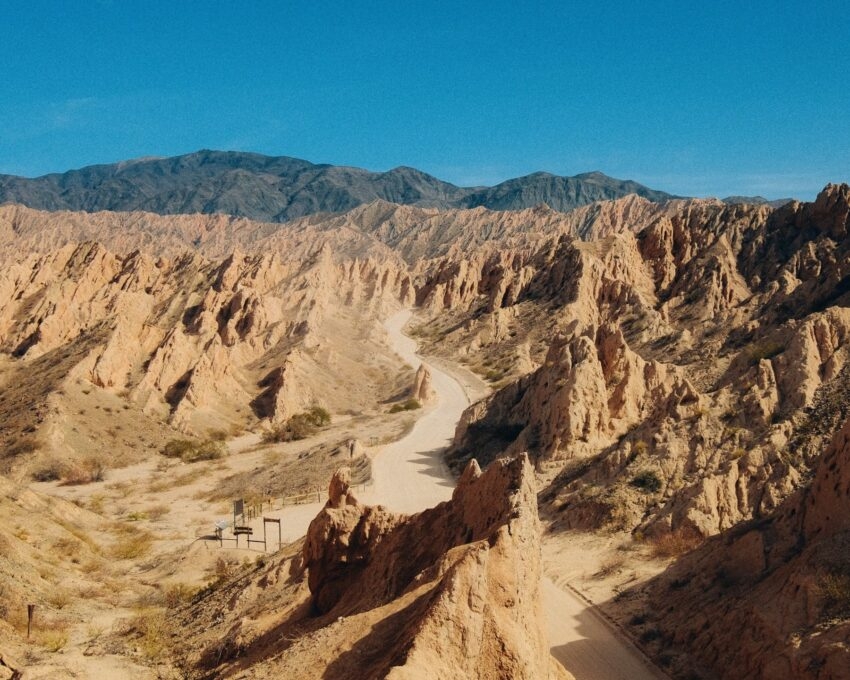 Narrow dirt road winding through sharp, angled sandstone rock formations in a desert canyon under a clear sky.