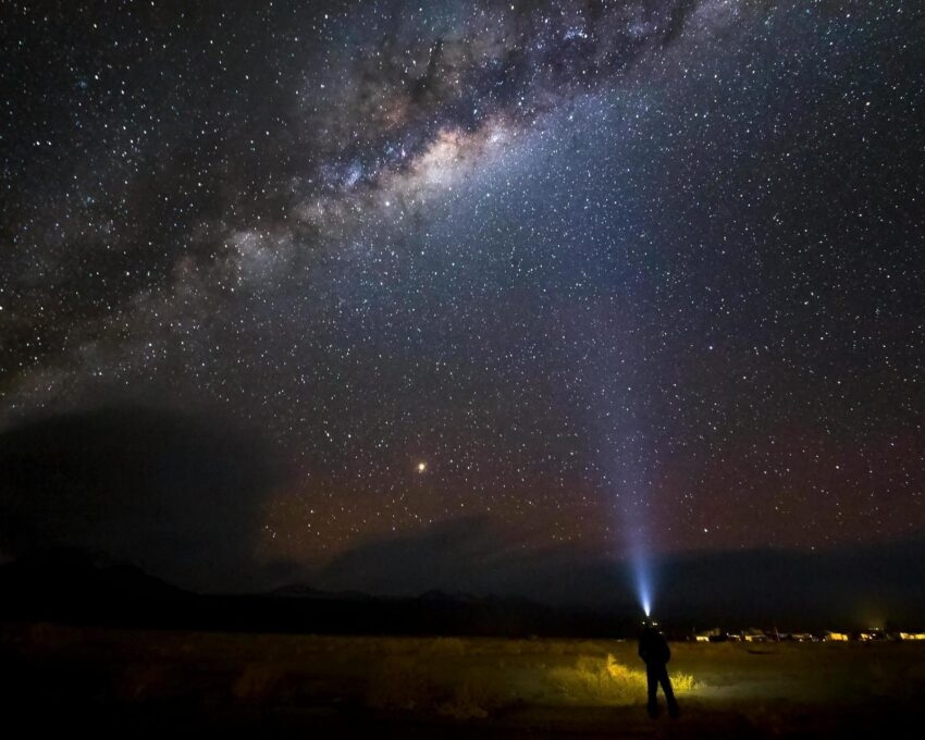 Silhouette of a person shining a light beam into a brilliant, star-filled night sky featuring the Milky Way galaxy.