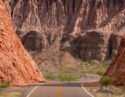 A winding road passes through towering red rock formations and desert shrubs under a bright sun.