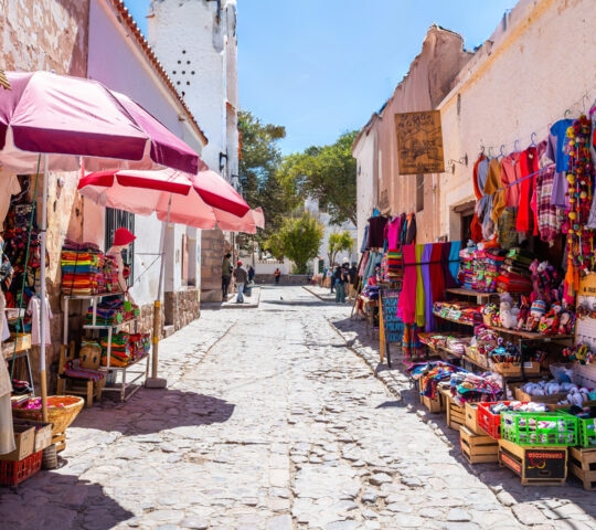 Narrow cobblestone street with market stalls displaying colorful t-shirts, blankets, and textiles.