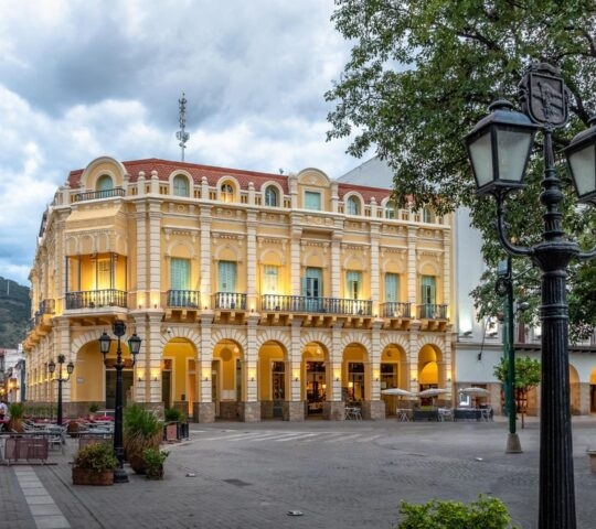 Illuminated yellow colonial building with ornate balconies and arched walkways in a city plaza at dusk.