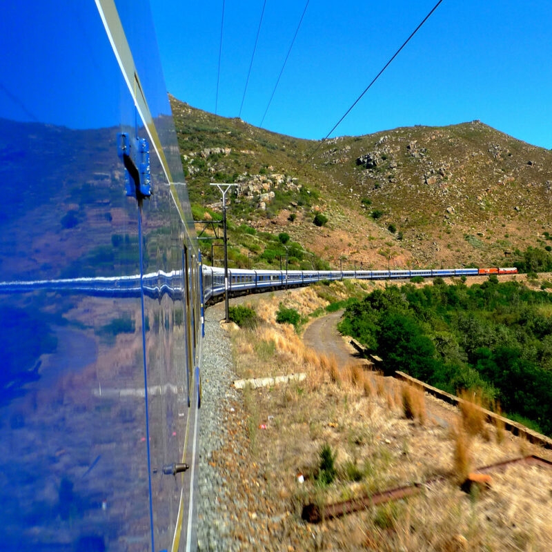 Reflection of a long luxury train on its blue side panels as it curves through a sunny mountain valley.
