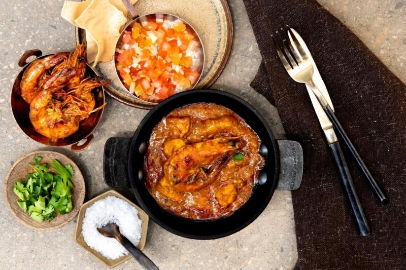 Top-down view of a prawn curry dish surrounded by small bowls of tomato salsa, salt, and fresh herbs.