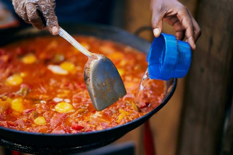 Close-up of hands cooking a red tomato and egg dish in a large black pan outdoors.