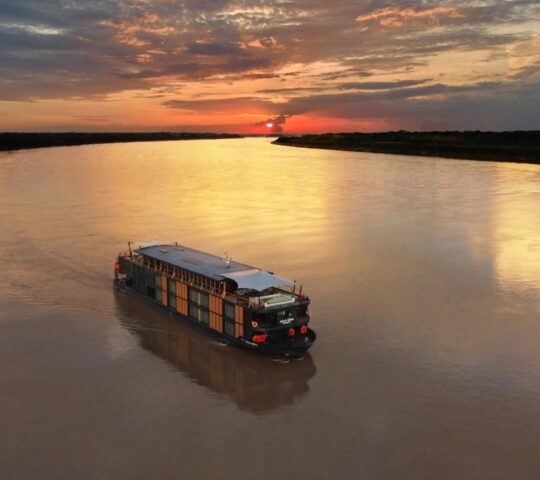 Aerial view of a luxury cruise ship on a wide river with a vibrant orange sunset reflecting on the water.