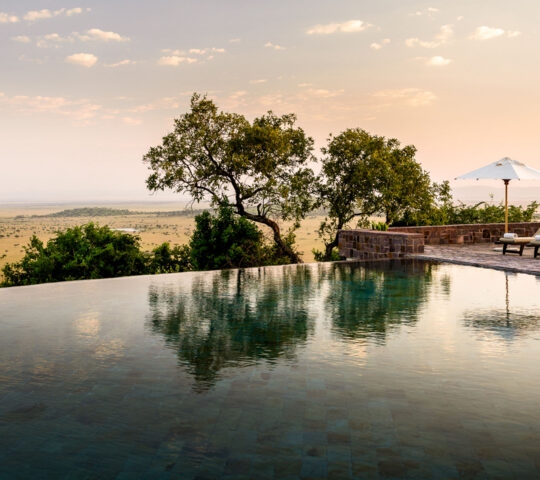 Infinity pool overlooking a wide grassy savanna under a pale sunset sky with lounge chairs and a white umbrella.