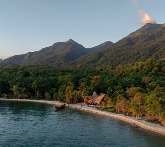 Aerial view of a remote beach lodge with thatched roofs on a white sand beach surrounded by dense jungle and mountains.