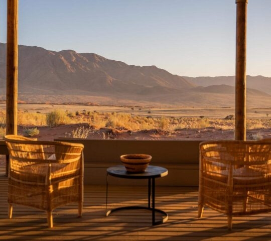Wicker chairs on a wooden terrace overlooking a desert valley and mountains at sunset.