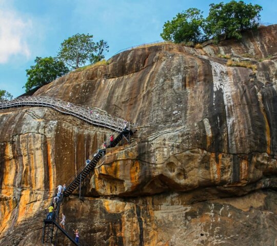 Visitors ascending a long metal staircase built into the side of a massive, weathered orange and grey rock.
