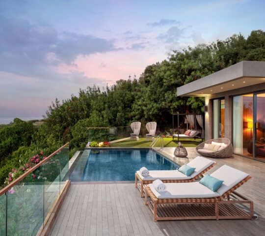 A private villa terrace with a swimming pool, wooden sun loungers, and lush greenery overlooking the sea at sunset.