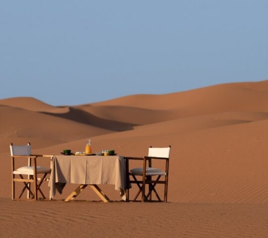 A breakfast table and two chairs set in the middle of vast desert sand dunes under a clear blue sky.