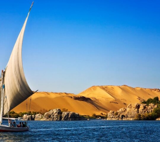 A white-sailed felucca boat on deep blue water with large sand dunes and rock formations in the background.