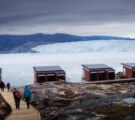 Four red wooden lodges at Eqi Glacier Lodge on a rocky cliff overlooking a glacier in Greenland