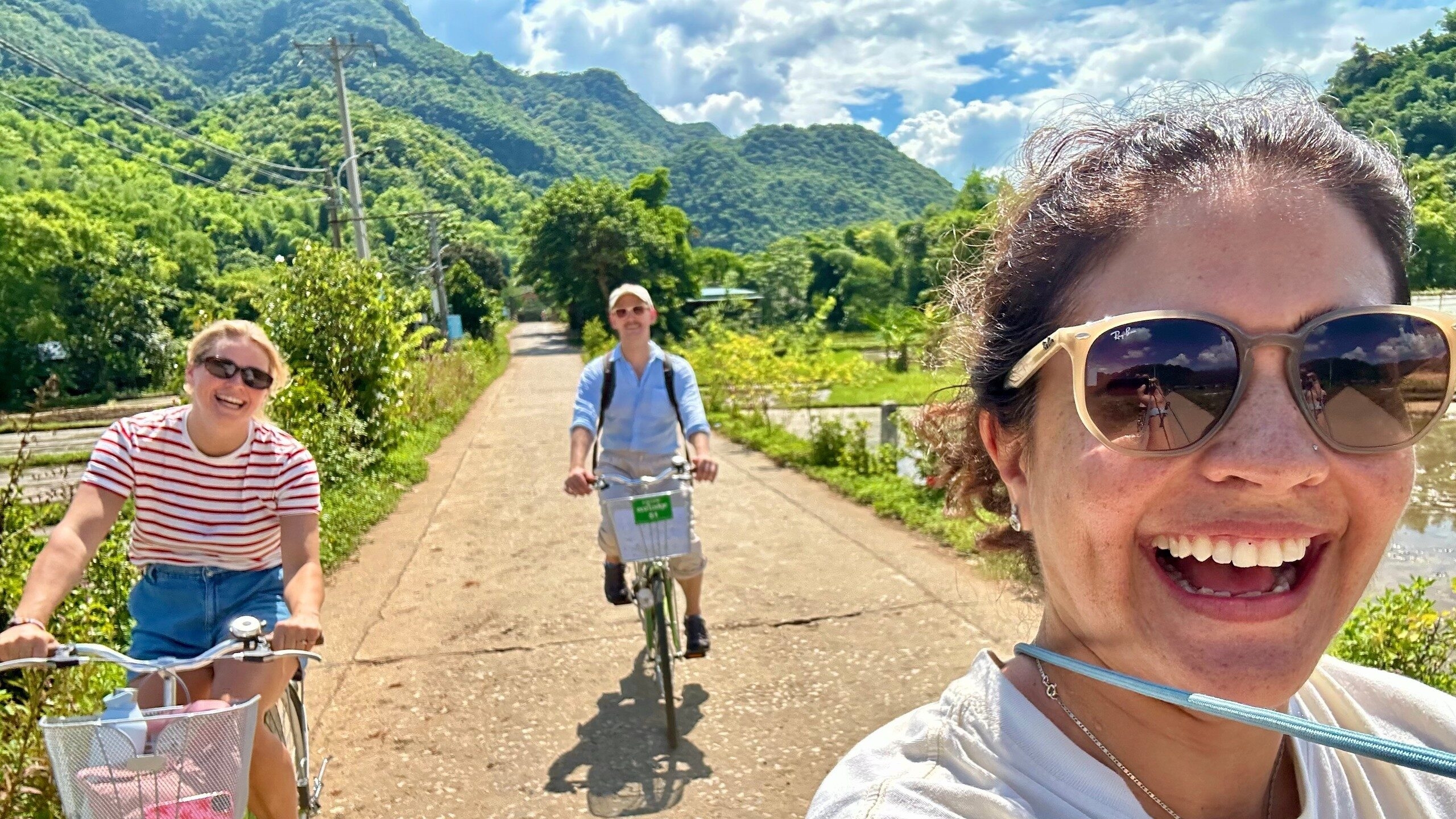 A group of three people smiling and laughing while riding bikes on a sunny path through lush green hills.