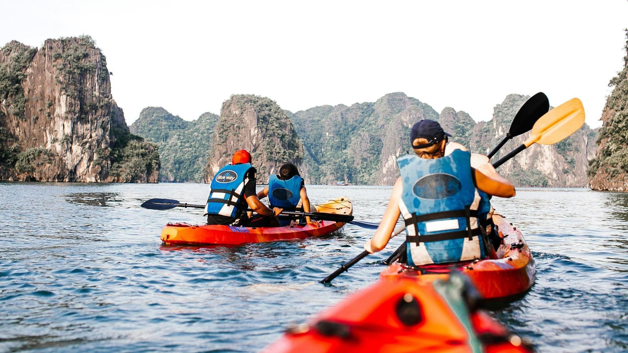 A line of red kayaks with people in blue life jackets paddling on a wide bay near rocky cliffs.