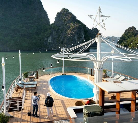 A cruise ship deck with a small oval pool, lounge chairs, and two people looking out at mountainous islands.