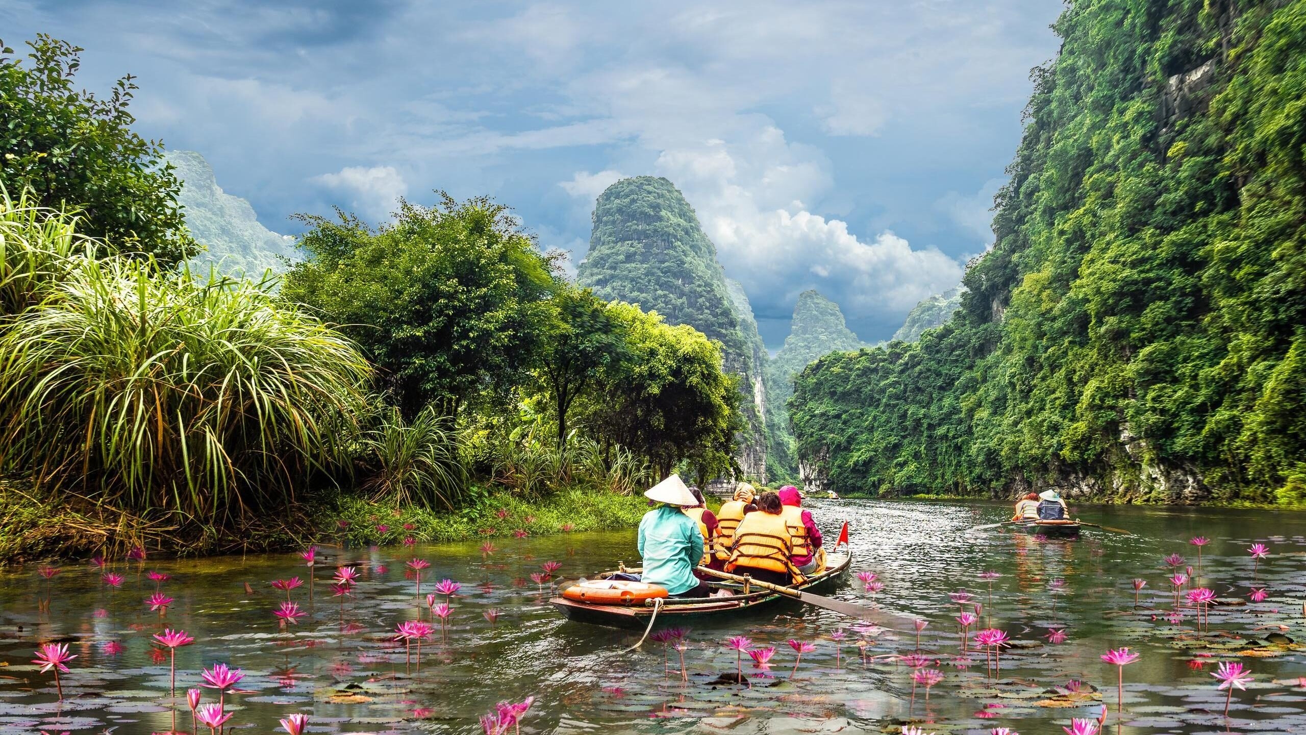 Several small boats rowing through a pond filled with pink lilies, surrounded by large green mountains.