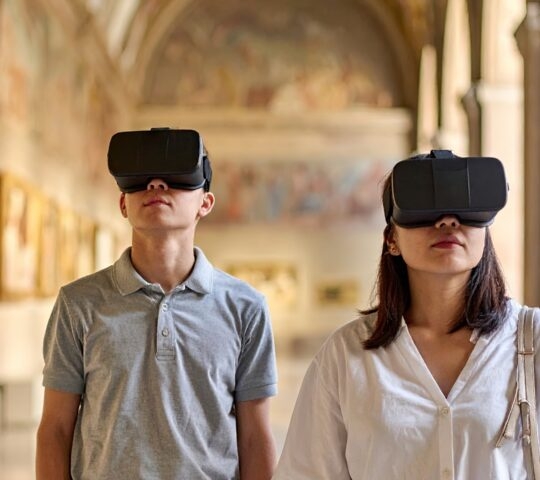 Two people wearing black VR goggles standing in a historic museum gallery with large wall paintings.