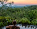 A woman in a plunge pool overlooking a vibrant jungle landscape as the sun sets in the horizon.