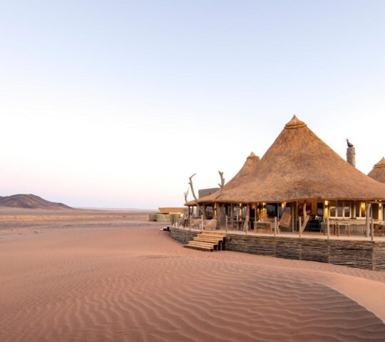 A thatched desert lodge with a wooden deck at Little Kulala Namibia, surrounded by rippled red sand dunes under a pale sky.