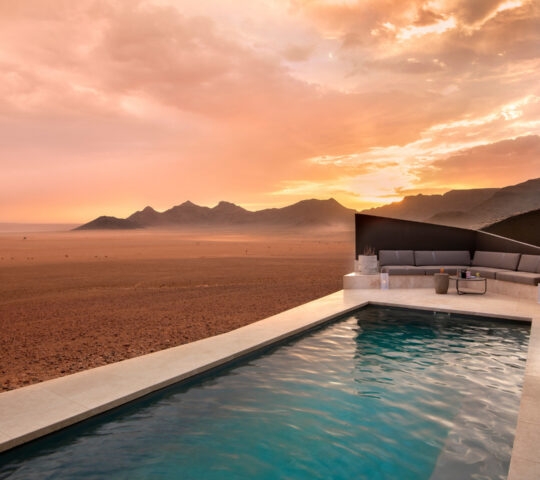 Private infinity pool and lounge area at Sossusvlei Desert Lodge overlooking a desert valley under a sunset sky.