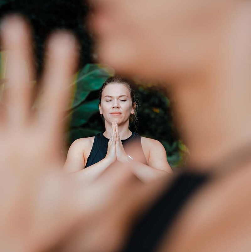 A woman closes her eyes during a yoga pose and looks peaceful with green foliage behind her and the yoga teacher in front of her.