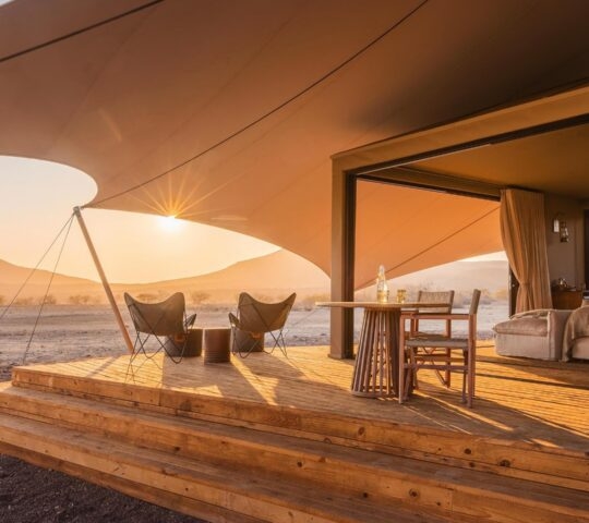 Tented guest suite at Wilderness Desert Rhino Camp with a wooden veranda, lounge chairs, and desert views at dawn.
