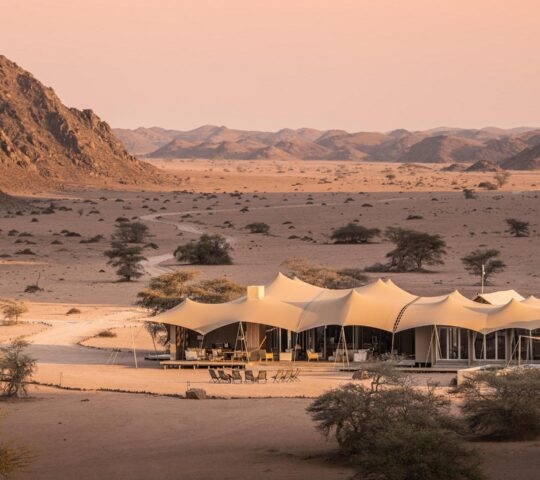Tented roofs of the luxury Hoanib Skeleton Coast Wilderness Camp in a vast desert valley surrounded by rocky hills.
