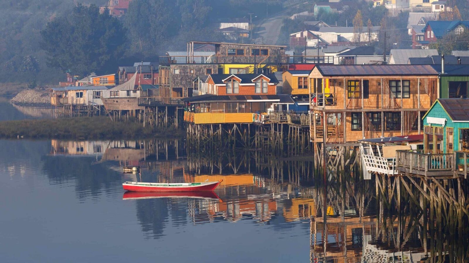 A red and white wooden boat on still water reflecting colorful houses built on stilts in the fog.