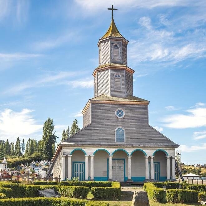 A three-story wooden church with a cross on top and blue accents on the ground floor arches on Chiloé Island.