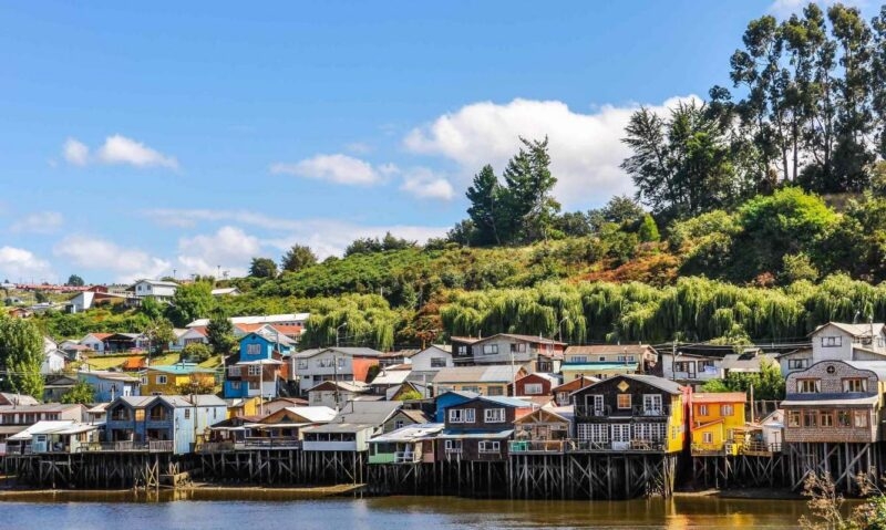 Brightly coloured wooden houses built on stilts over the water on Chiloé Island, Chile, against a green hillside.