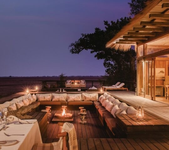 Sunken outdoor lounge with fire pit and dining table on a wooden deck overlooking the Zambian savanna at dusk.