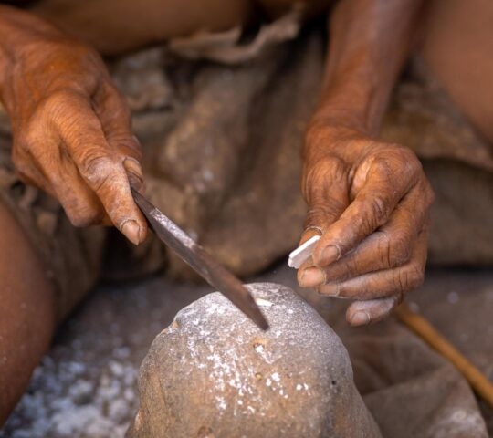 Close-up of hands using a metal tool and a stone to craft and shape small white fragments on a workspace.