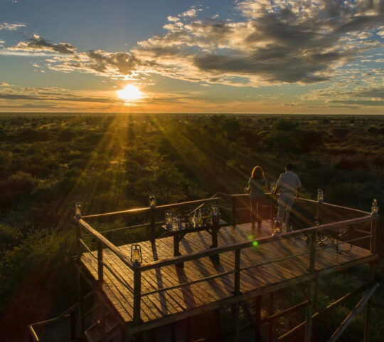 Two people standing on a wooden lookout platform at sunset, overlooking a vast savanna landscape.