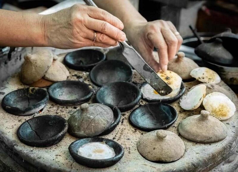 Hands using a spatula to cook small eggs in individual circular molds of a traditional clay griddle with ceramic lids.