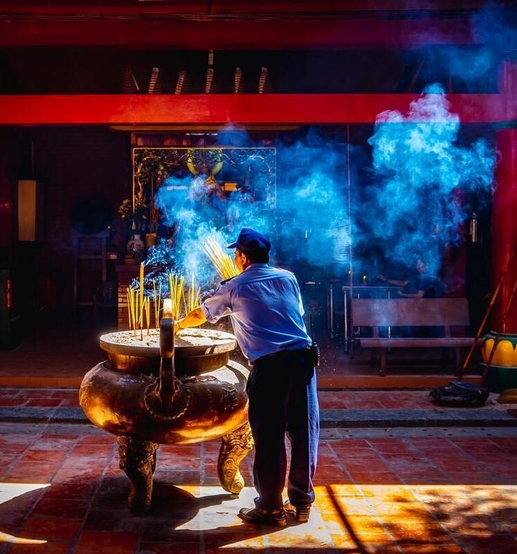 A man stands before a large ornate incense burner as shafts of light illuminate thick plumes of blue smoke in a temple.