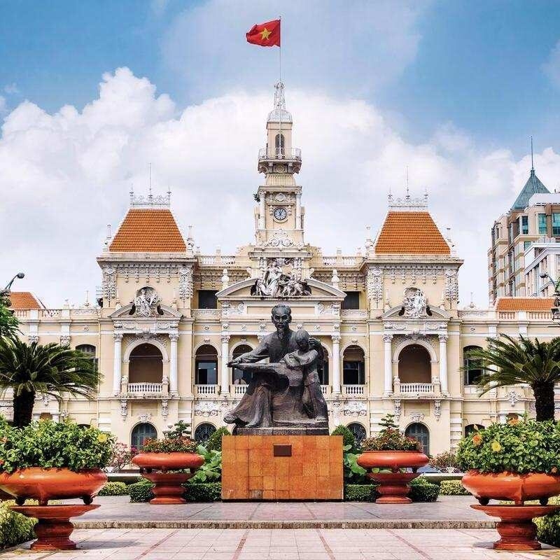 The grand, light-yellow Ho Chi Minh's city hall building with a central clock tower and a statue situated in a manicured courtyard.