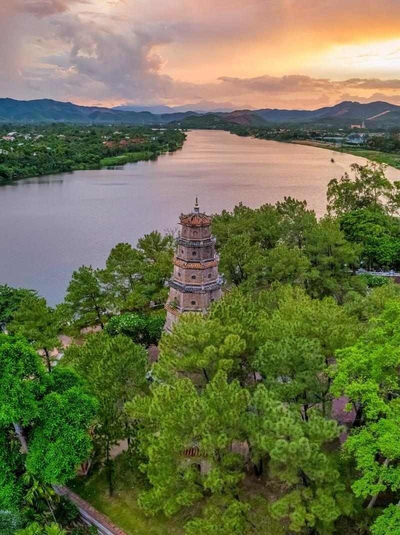 High-angle view of a tall octagonal pagoda surrounded by lush trees next to a wide river during a golden sunset.
