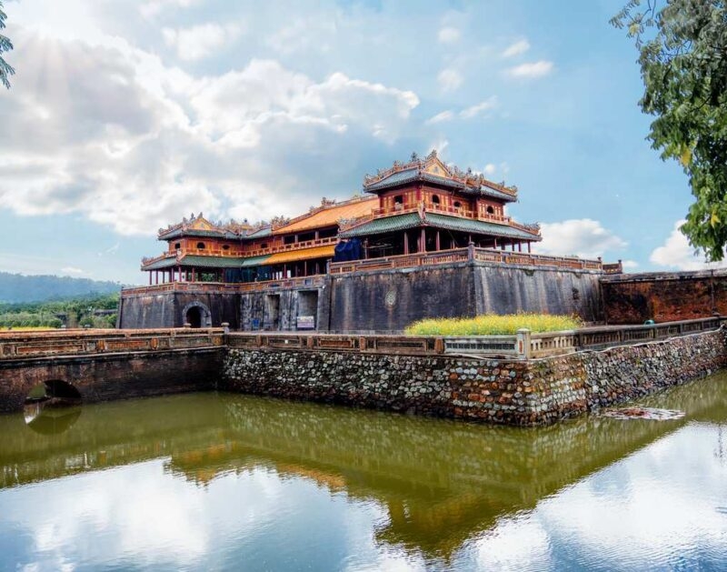 A traditional Vietnamese palace with yellow walls and tiered roofs sits behind a stone moat under a cloudy blue sky.