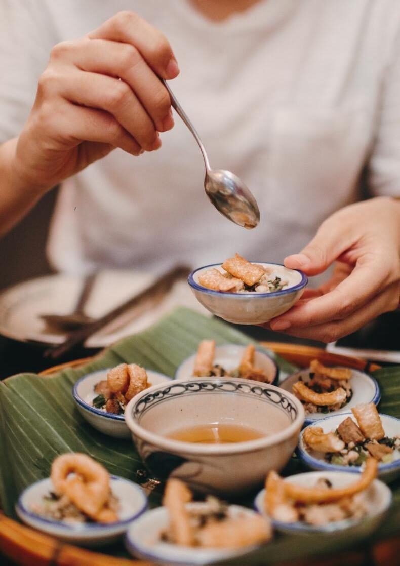 Close-up of a person holding a small ceramic bowl of rice cakes topped with crispy pork skin and scallions.
