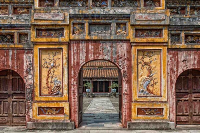 An ornate, weathered stone archway with decorative panels showing birds and trees leads into a courtyard with a temple.