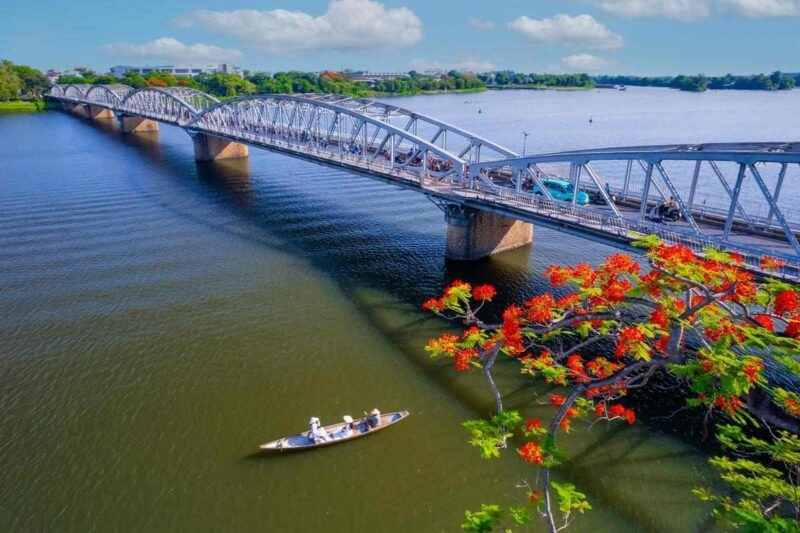 A high-angle view of a long arched bridge spanning a wide river with a small boat and red flowering trees in the foreground.
