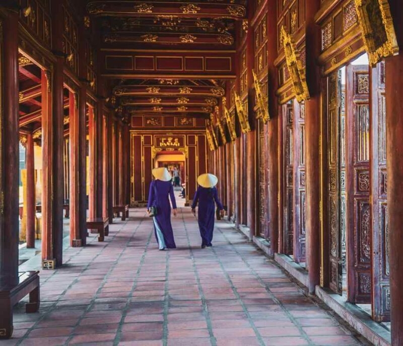 Two women in purple dresses and hats walk down a long, ornate wooden corridor with red columns and gold carvings.