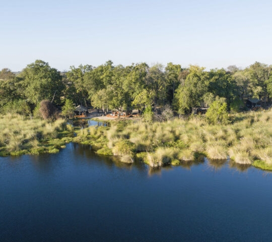 High angle view of a safari camp hidden in trees next to a blue waterway and grassy marshland.